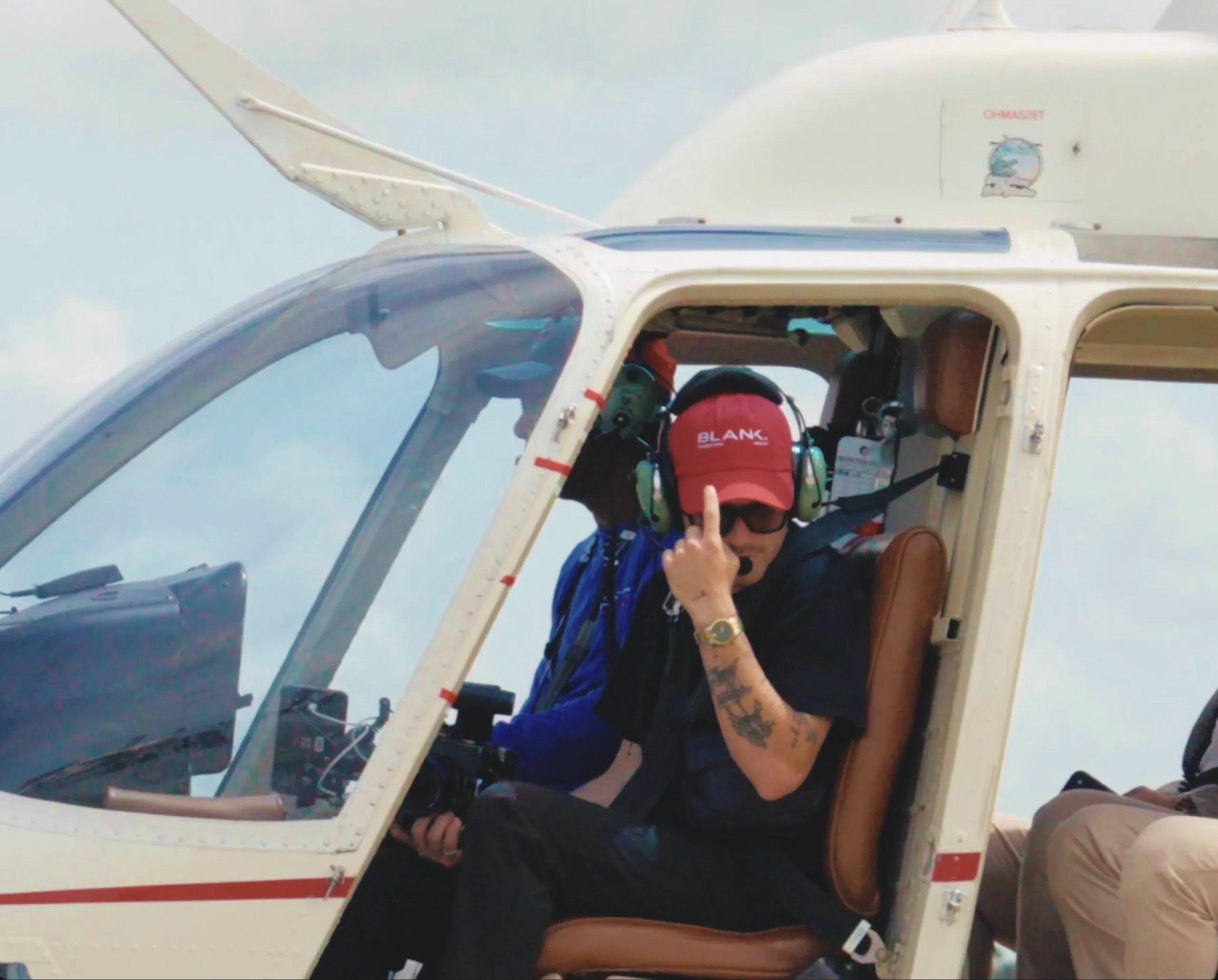 Behind the scenes — crew member in a Blank cap seated in a helicopter before an aerial shoot.