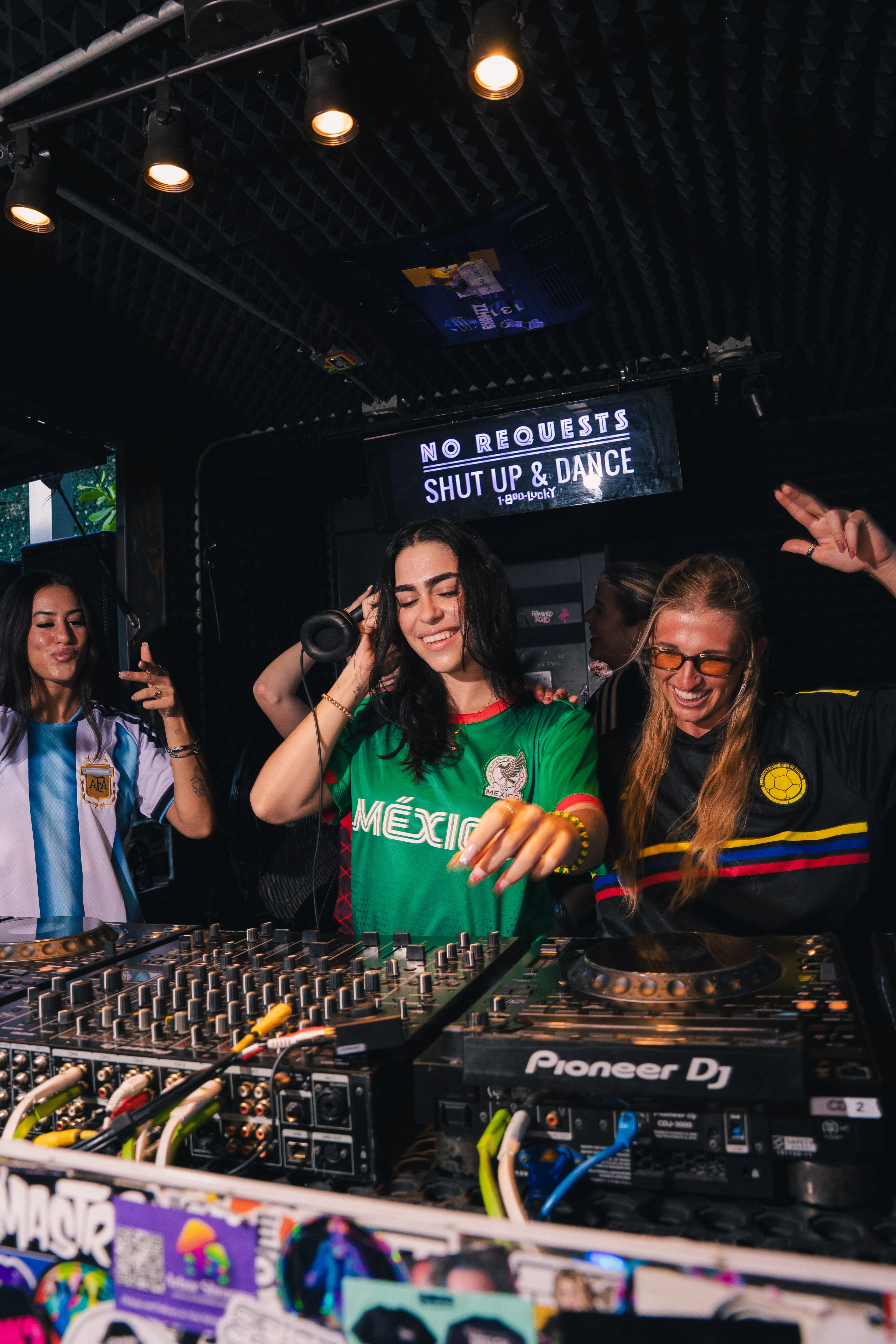 A World Cup-season night at 1-800-Lucky — a DJ in a green Mexico jersey mixes on Pioneer decks between friends in Argentina and Colombia jerseys, under the club's 'No Requests, Shut Up & Dance' sign.