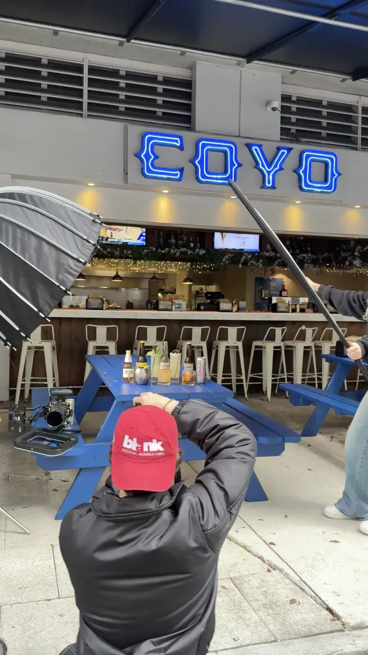 A crew member in a red cap films the blue neon COYO storefront and its bar stools from behind a boom, an umbrella rig in frame.