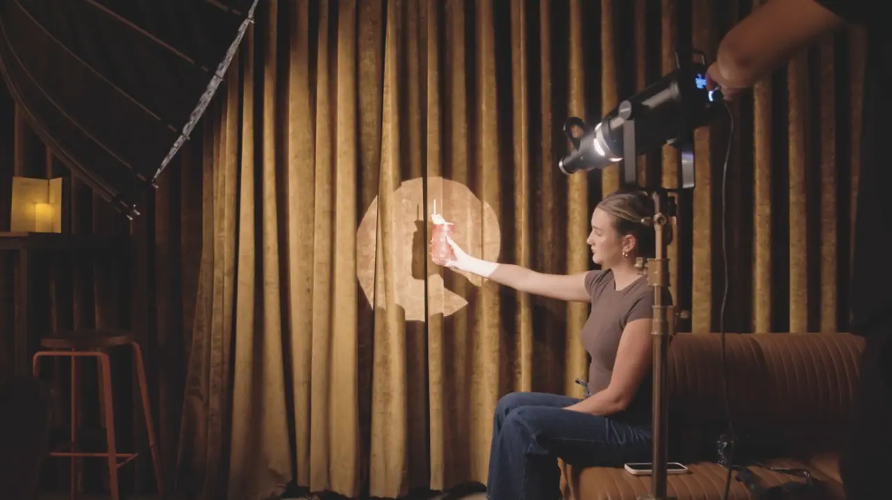 Behind the scenes at a Dante's HiFi+ shoot — a stylist holds a cocktail toward a cinema light amid softboxes and camera rigs on set.