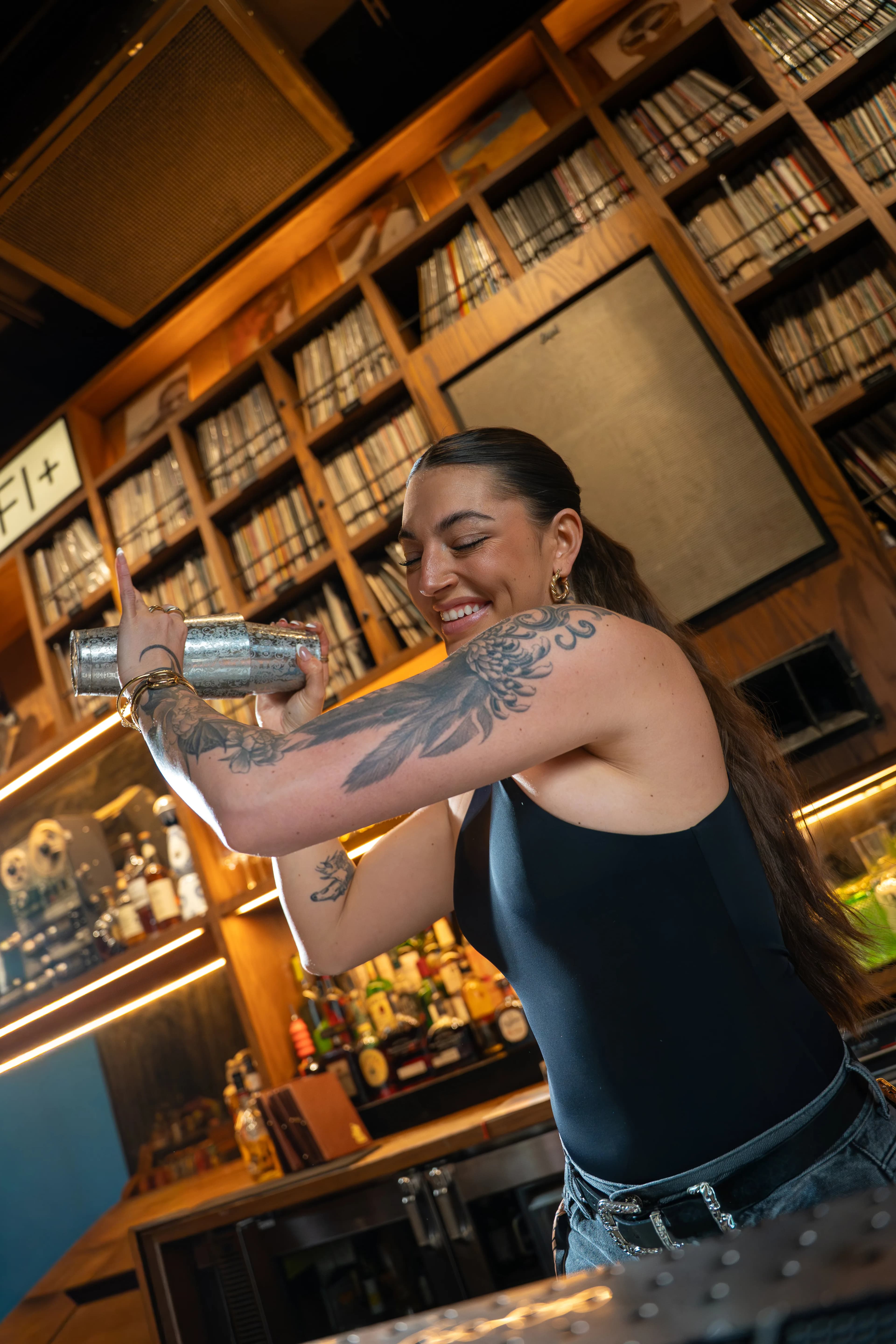 A smiling bartender with a tattooed arm shakes a cocktail tin in front of a floor-to-ceiling wall of vinyl records at Dante's HiFi+.