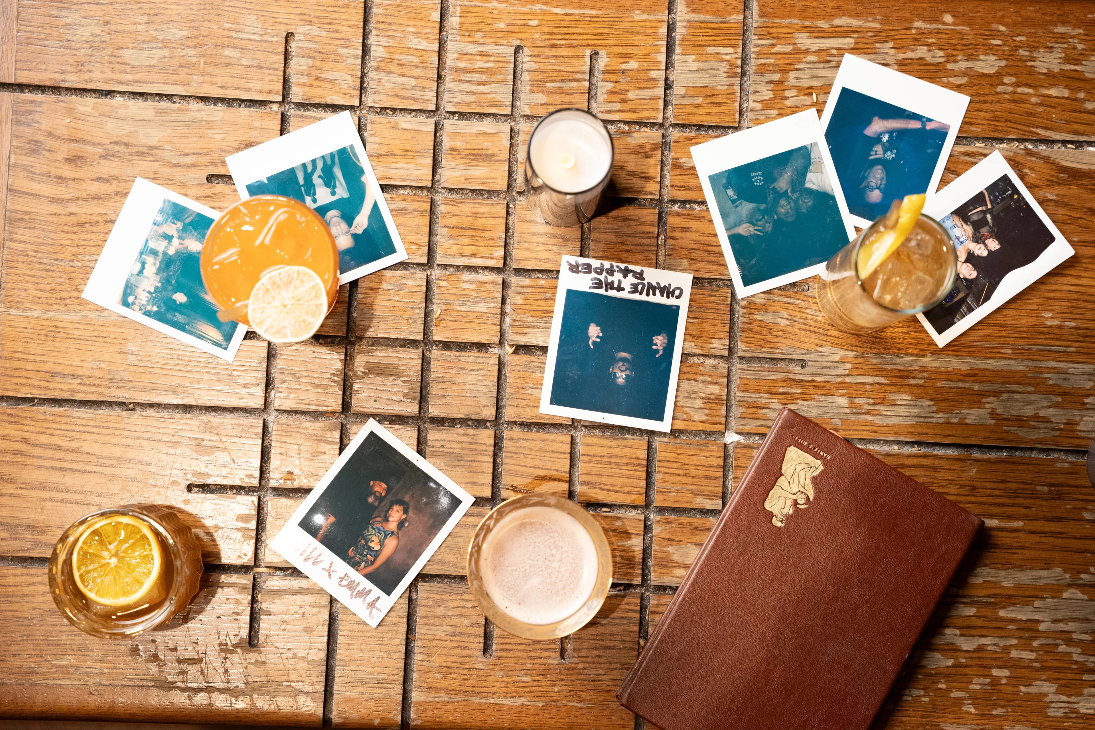 A top-down flat-lay of scattered polaroid snapshots of nightlife moments among cocktails, a candle, and a leather-bound book on a weathered wood table at Dante's HiFi+.