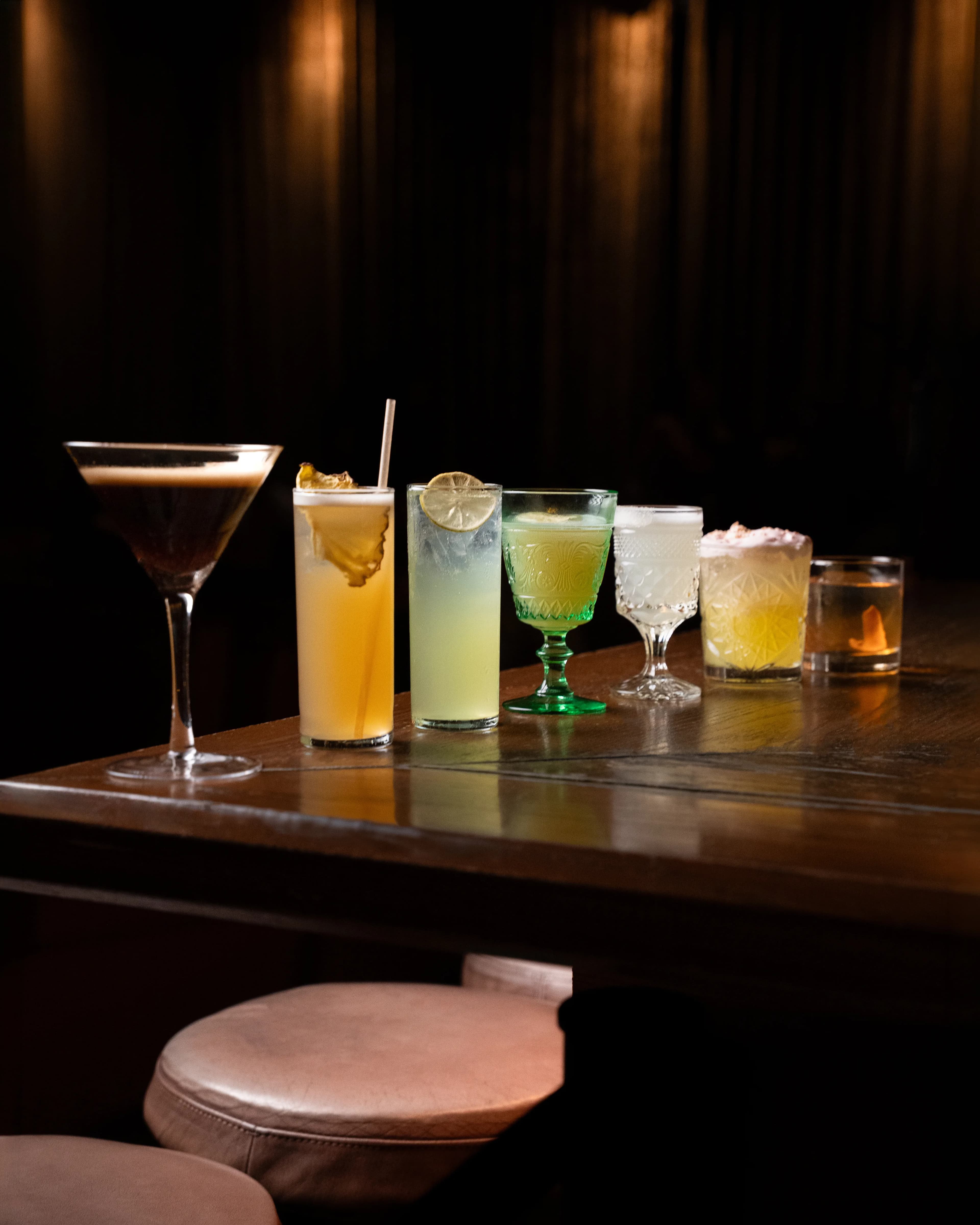 A row of seven cocktails in varied glassware lined up along a polished wood bar in the low light at Dante's HiFi+.