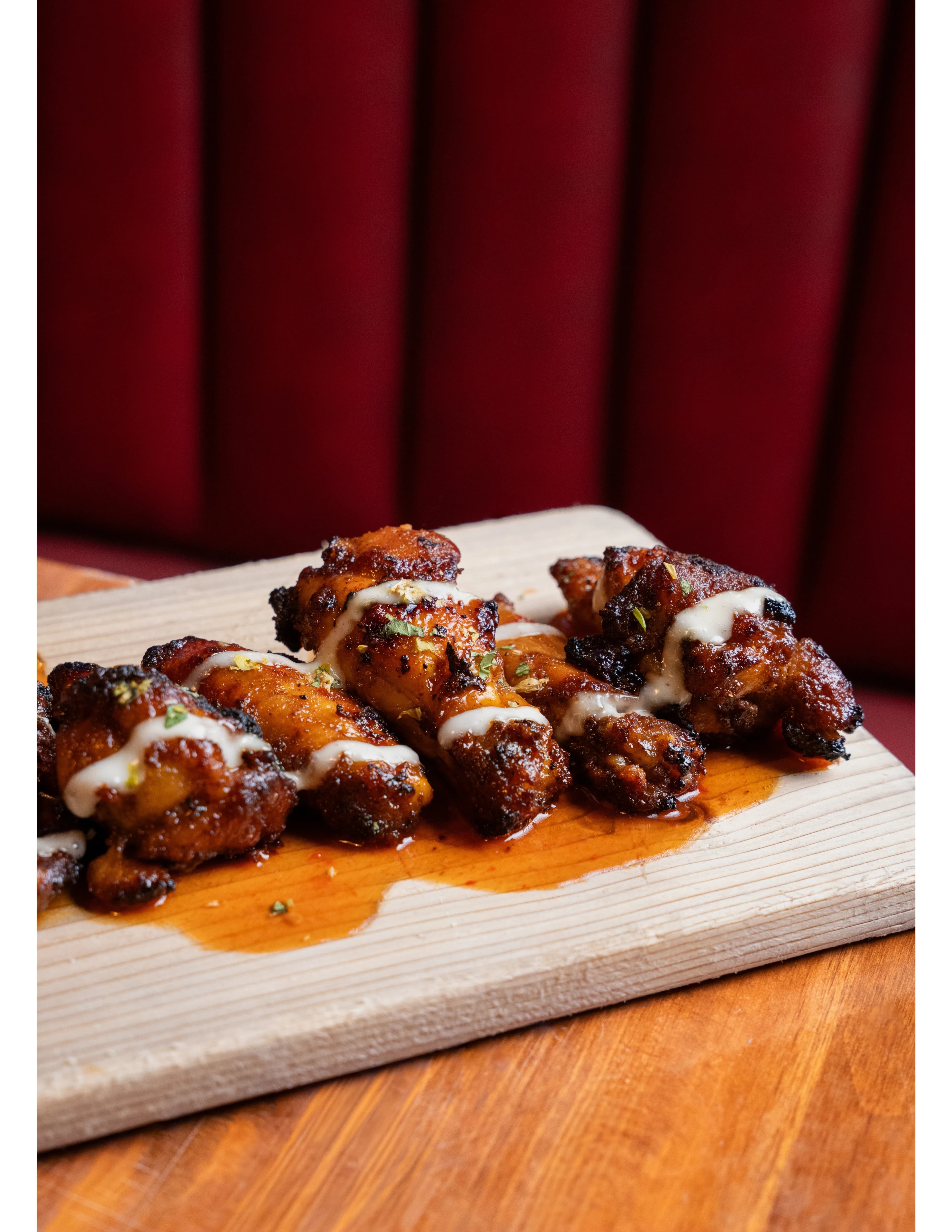 Glazed, herb-flecked chicken wings drizzled with white sauce on a wood board against a red velvet booth at DC Pie Co.