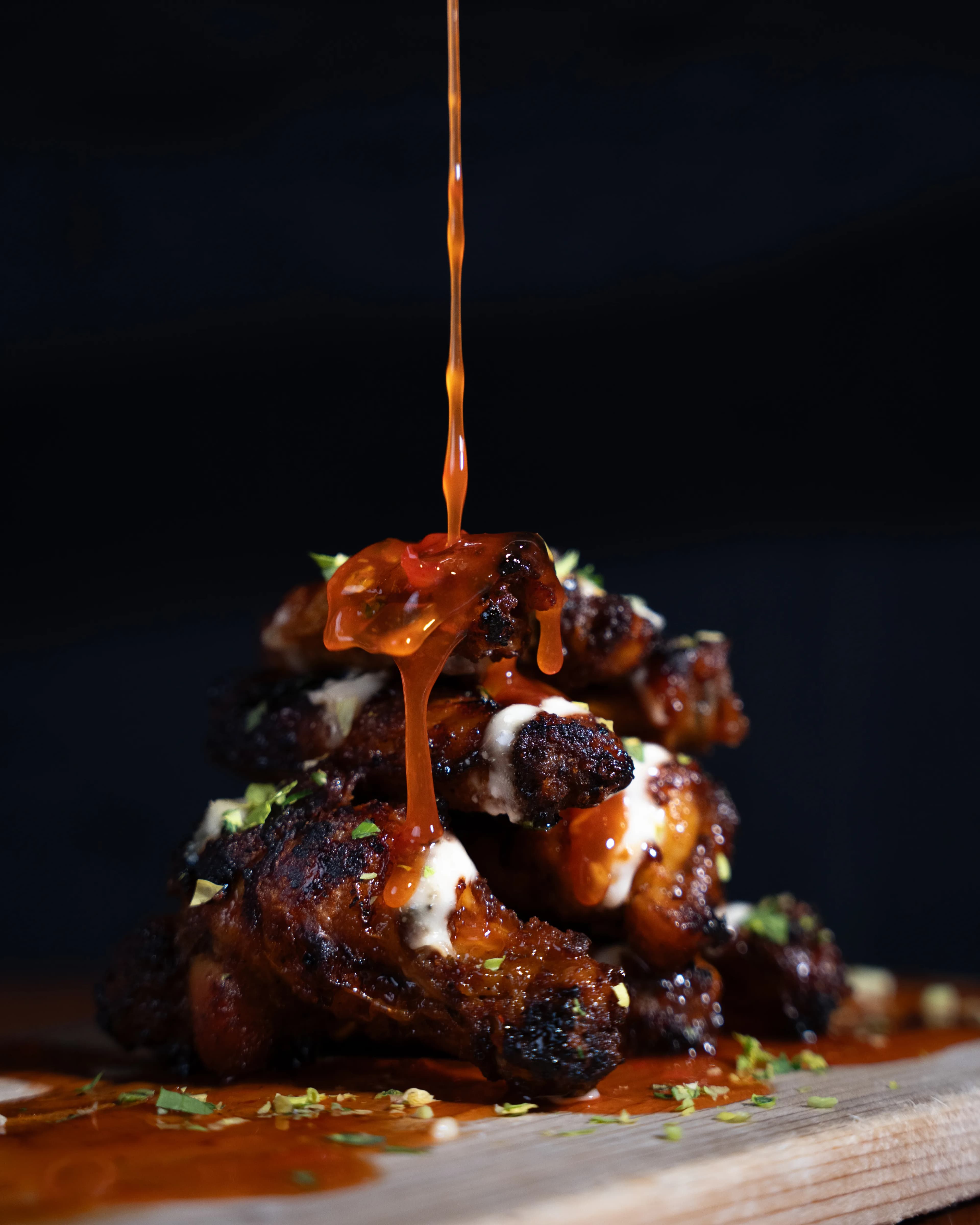 Hot sauce pours over a stack of charred wings dressed with white drizzle in moody light at DC Pie Co.