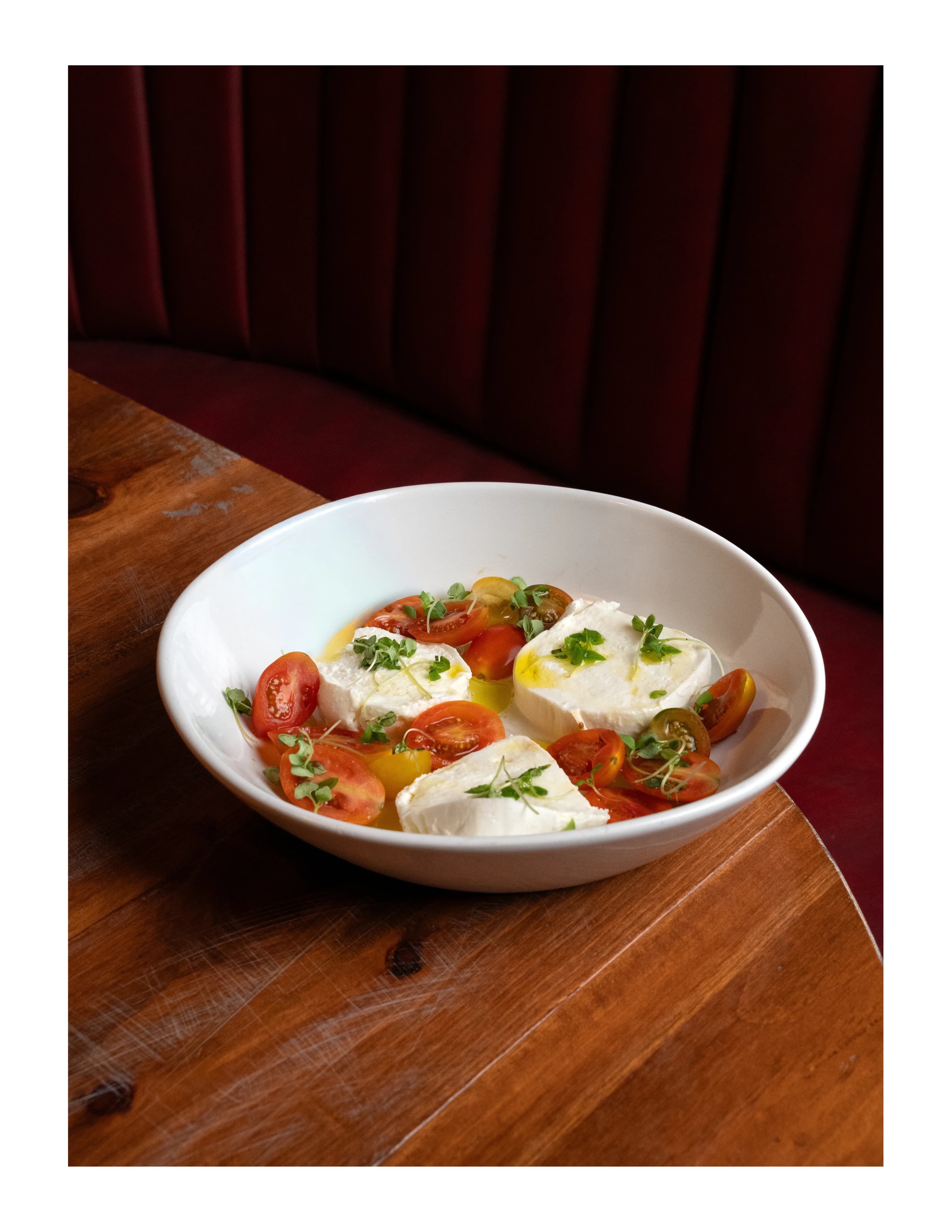 Fresh mozzarella with cherry tomatoes and herbs in a white bowl on a wood table against red velvet at DC Pie Co.