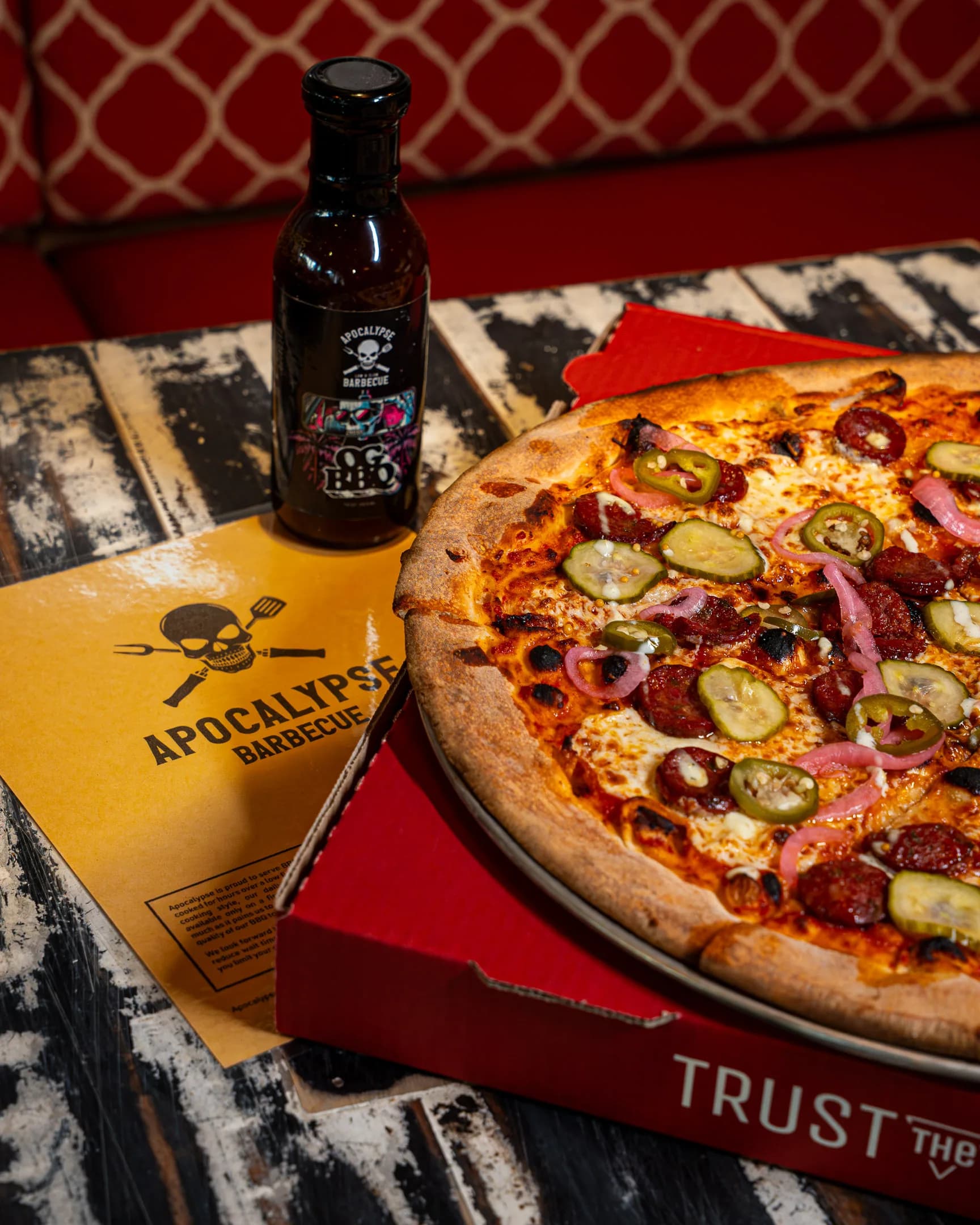 A loaded DC Pie Co. pizza with pepperoni and jalapeños beside an Apocalypse BBQ hot sauce bottle on a red 'Trust the Crust' box.