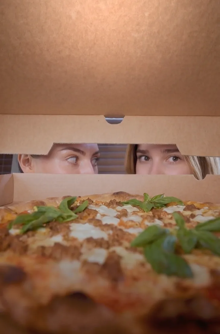 Two guests peer into an open pizza box at an arugula-topped DC Pie Co. pizza.