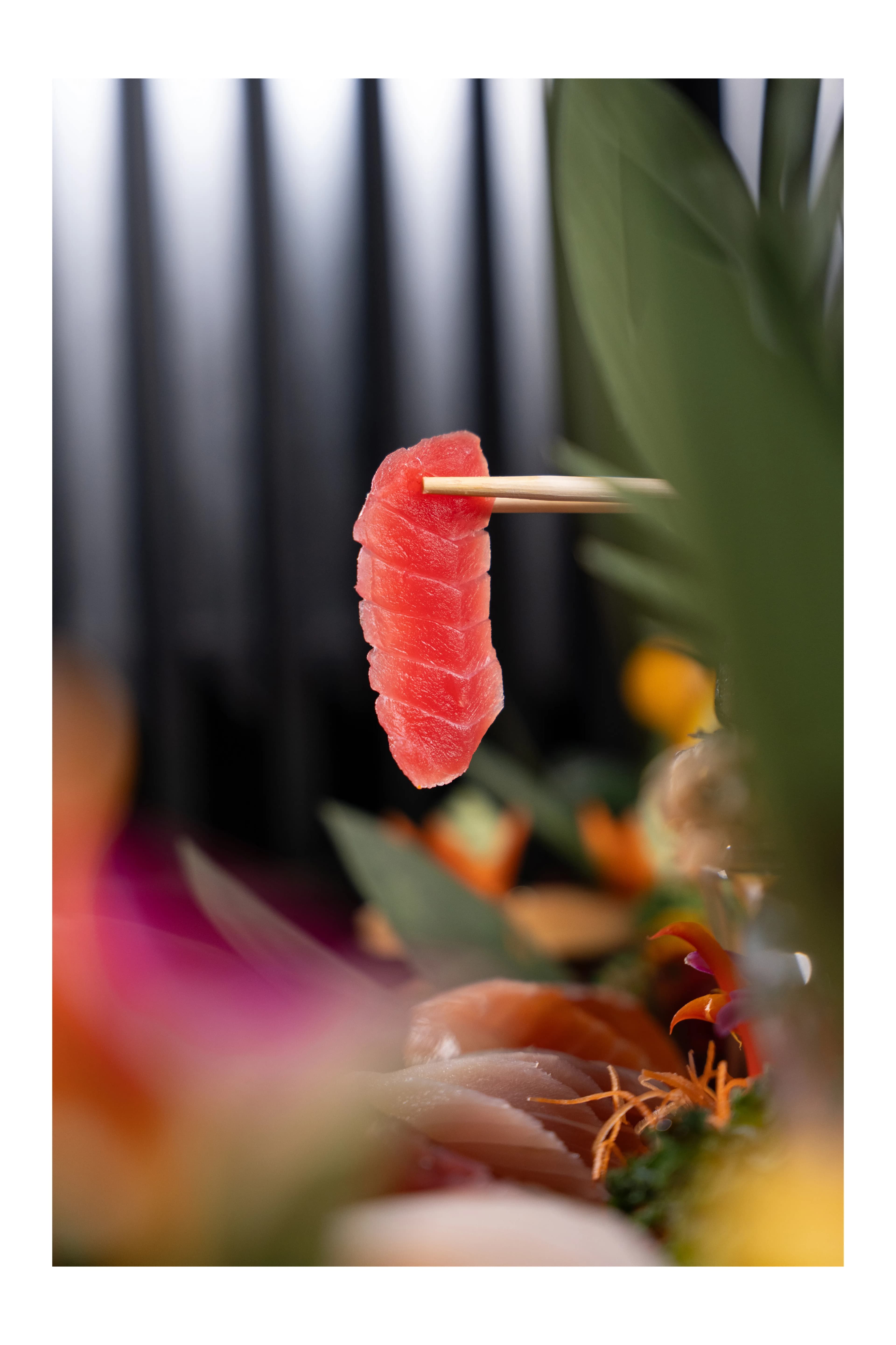 A slice of ruby tuna held upright on a skewer against a soft-focus floral arrangement at Sushi Sake.