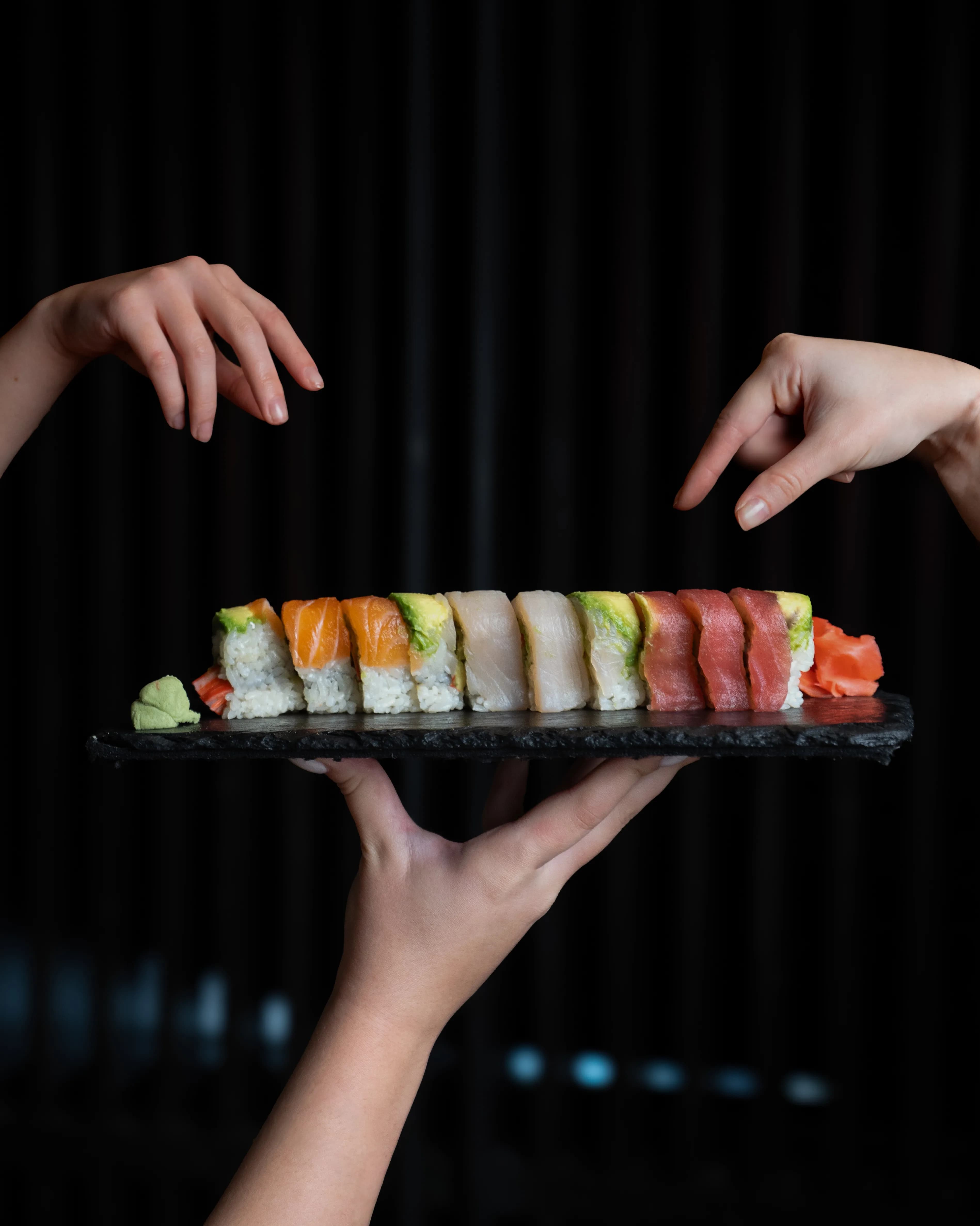 Hands present a long tray of colorful nigiri and rolls in low light at Sushi Sake.