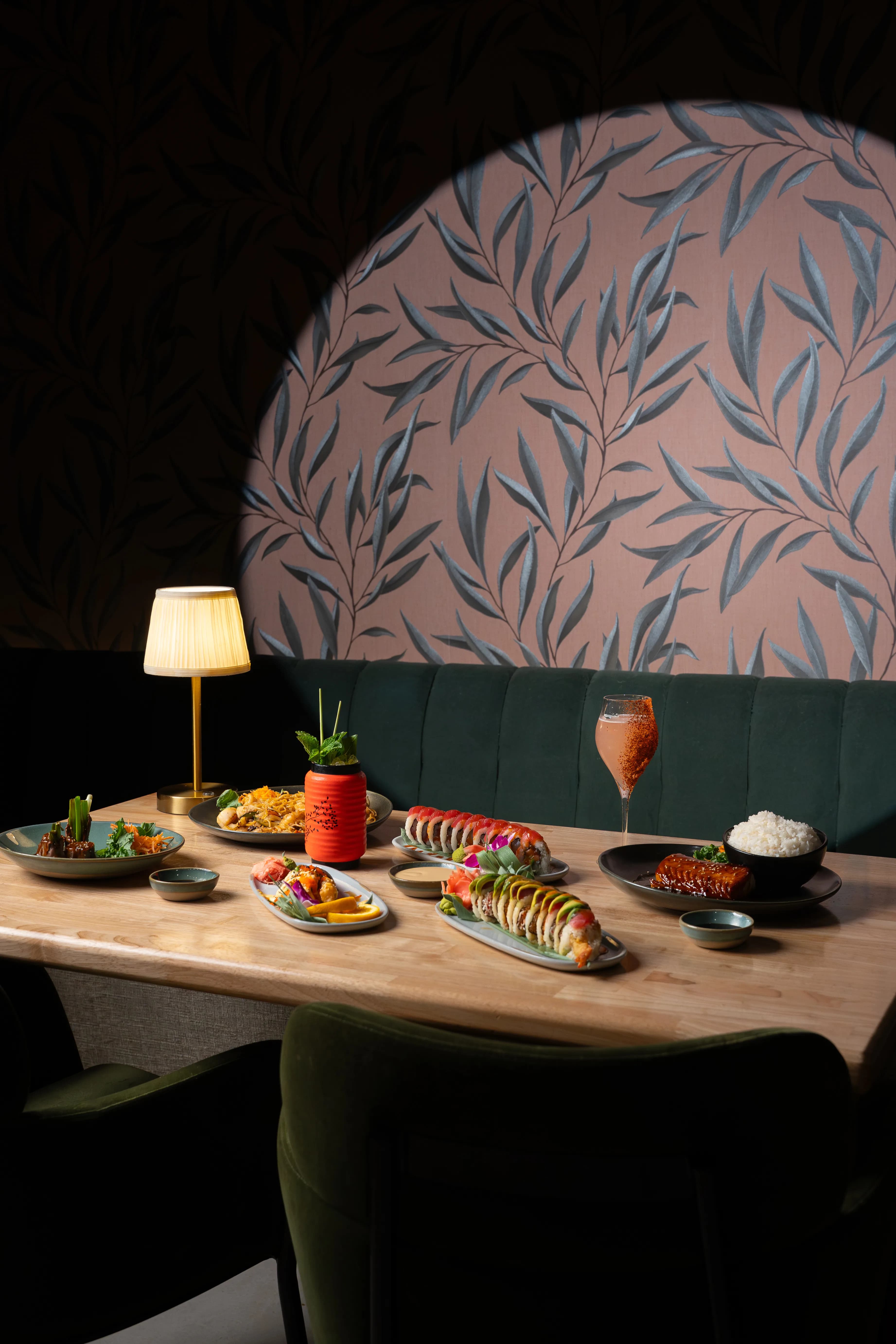 A styled table of dishes against a pink botanical booth with a warm lamp at Sushi Sake.