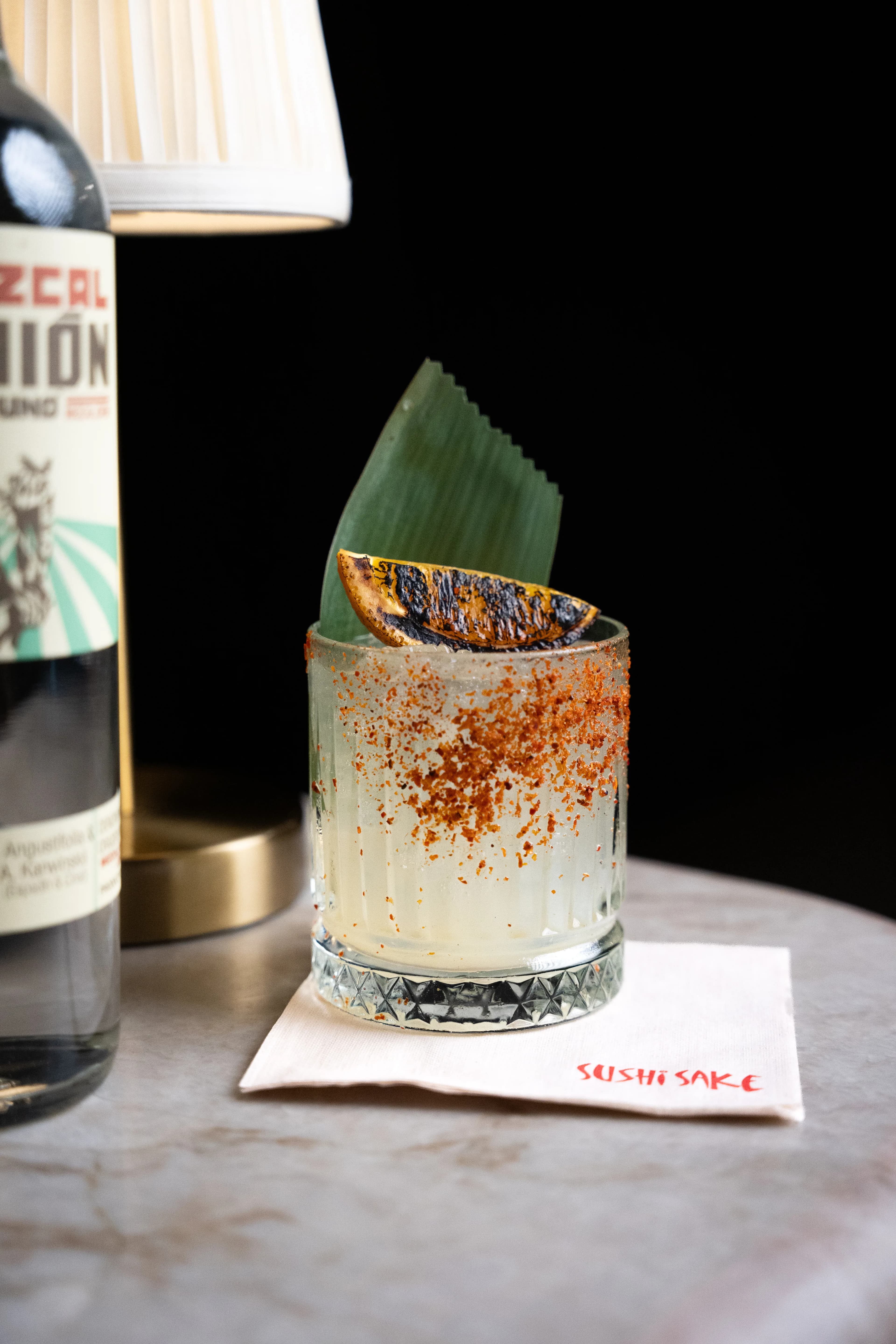A garnished Sushi Sake cocktail with a shiso leaf and chili rim beside a bottle.