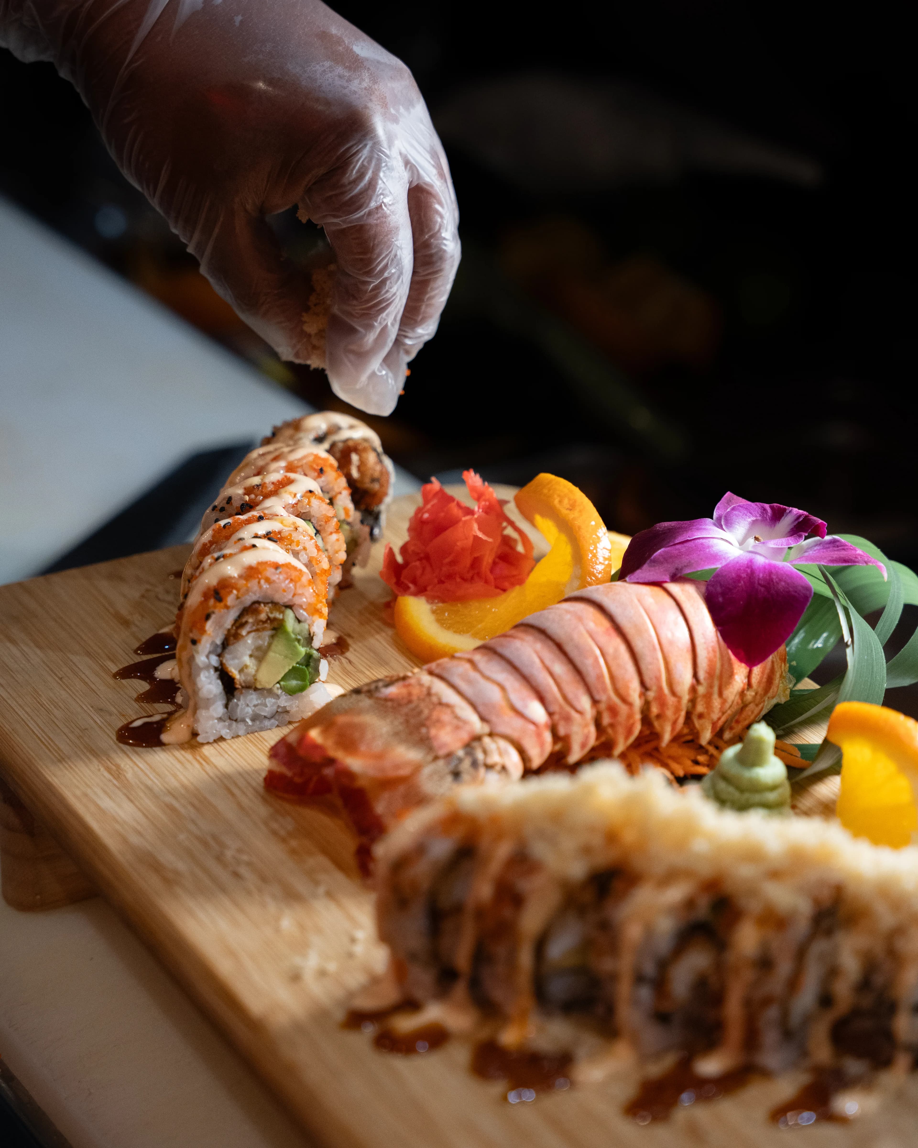 A gloved hand plates a Sushi Sake specialty roll with lobster and an orchid on a wood board.